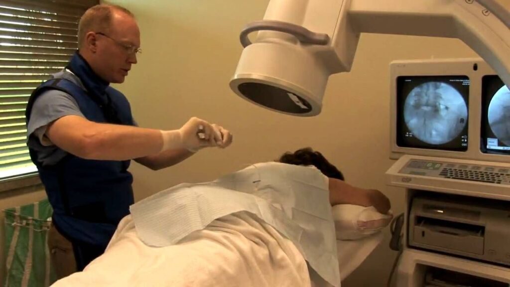 A medical professional in protective gear performs a spinal injection or procedure on a patient lying face down under a fluoroscopy machine. The procedure is being guided by live X-ray images displayed on two monitors beside the patient.
