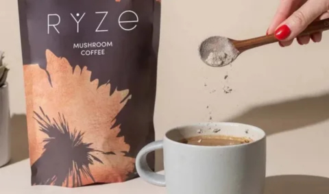 Hand sprinkling mushroom coffee powder into a mug beside a Ryze mushroom coffee package.