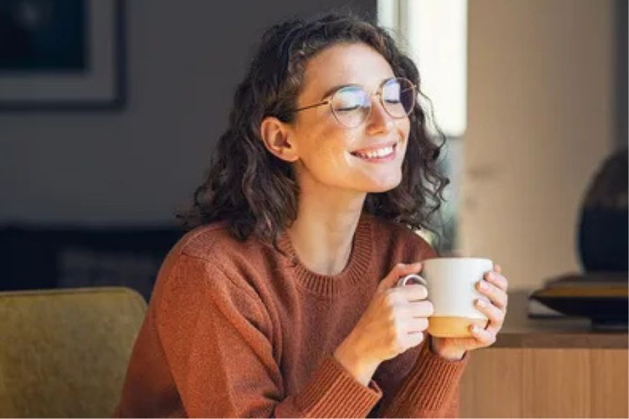 "A smiling woman in glasses sits indoors holding a warm cup of coffee, symbolizing the cautious return to coffee after vasectomy recovery."