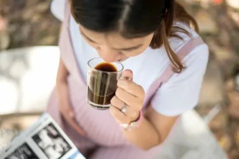 "Pregnant woman in pink overalls holding a glass mug of hot drink while reading, illustrating safe ways to enjoy hot chocolate during pregnancy."