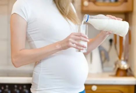 "Pregnant woman in white shirt pouring a glass of milk in the kitchen, symbolizing safe daily nutrition and moderation when drinking Ensure during pregnancy."