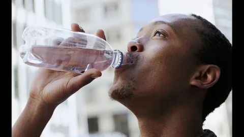 A close-up of a man drinking water from a clear plastic bottle outdoors, illustrating the act of hydrating to combat the effects of allergies during high-pollen seasons.