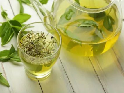 A close-up of a glass teapot and a small glass of light green herbal tea on a white table, illustrating the relaxing beverage often consumed in the evening.