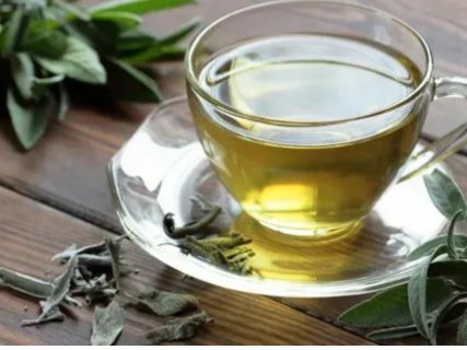 A glass teacup of green herbal tea on a wooden table, surrounded by loose leaves, symbolizing the relaxing, antioxidant-rich beverage recommended for nighttime consumption.
