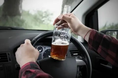 A person's hands on a steering wheel, with one hand holding an open bottle of alcohol in the car's passenger area, illustrating an open container violation.