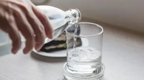 A hand pouring water slowly from a bottle into a clear glass on a coaster, illustrating the advice to sip water slowly and ensure a clean source to avoid or stop water-induced nausea.