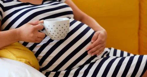 A pregnant woman wearing a striped dress, sitting and gently touching her belly while holding a mug of coffee or tea, symbolizing safe, moderate caffeine consumption during pregnancy.
