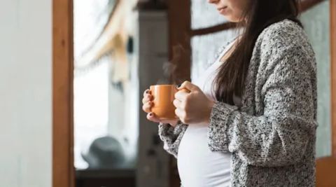 A side profile of a pregnant woman standing indoors, holding a steaming mug of coffee, illustrating the common belief or question about whether coffee can induce labor.