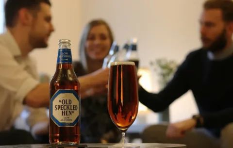 "A bottle and glass of ""Old Speckled Hen"" low-alcohol beer in the foreground, with blurred figures clinking glasses in the background, symbolizing the modern, balanced approach of moderation that many Quaker groups now take toward alcoholic drinks."