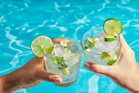 Two people clinking glasses filled with ice, lime, and clear drinks beside a bright blue pool, representing safe hydration options for travelers concerned about drinking water in Cancun.