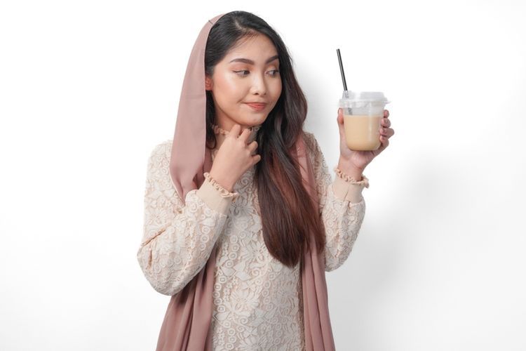 A Muslim woman holding a cup of iced coffee with a thoughtful expression, symbolizing questions about whether Muslims can drink coffee during fasting in Ramadan.