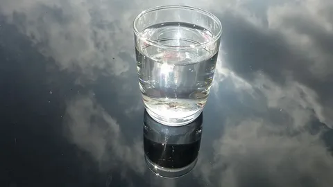 Glass of collected rainwater on reflective surface — illustrates that rainwater can appear clear but may contain airborne pollutants, bacteria, PFAS and microplastics, so filter or boil before drinking.